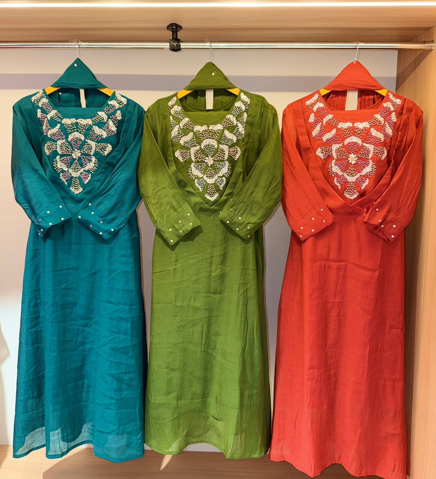 Three dresses in teal, green, and red with intricate lace details on a rack.